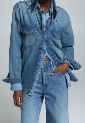 Person iført lagdelt blå denimjakke og skjorte med matchende blå denimjeans, den ene hånd i lommen, beskåret under skuldrene.