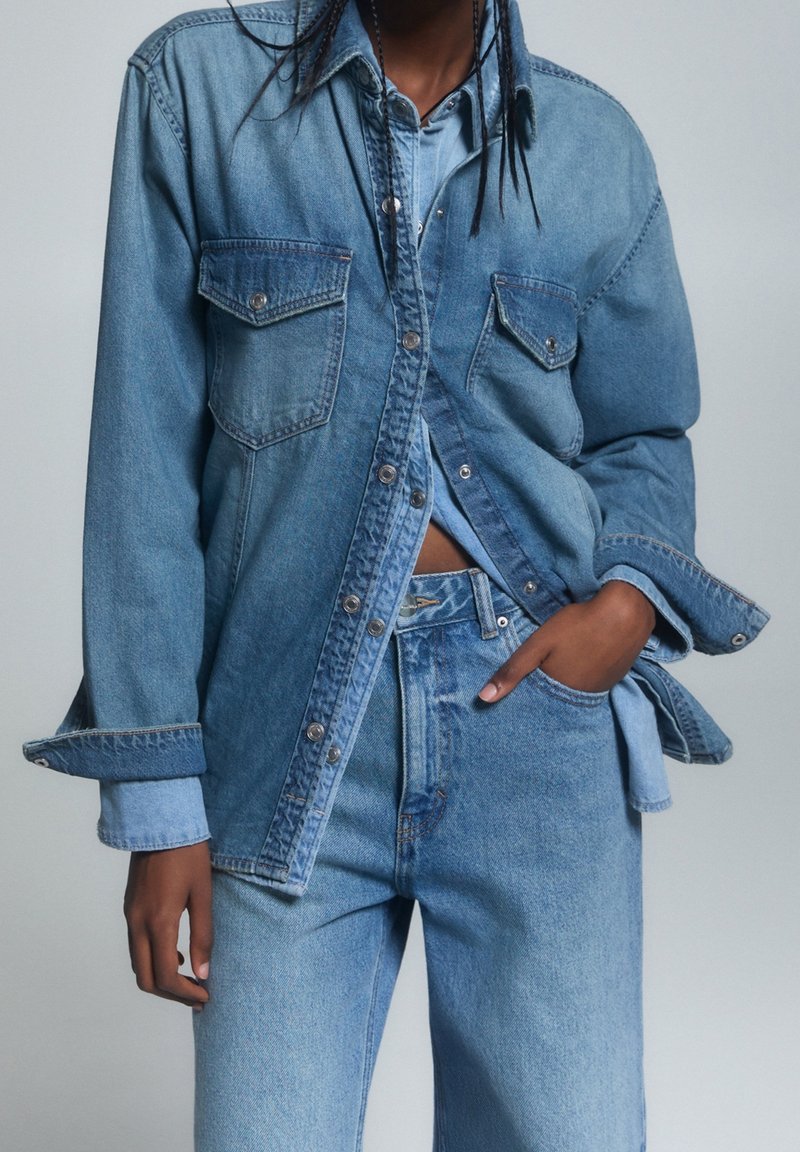 Personne portant une veste en denim bleu superposée et une chemise avec un jean en denim bleu assorti, une main dans la poche, cadrée sous les épaules.