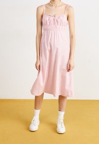 Robe rose sans manches avec un corsage froncé et un détail en ruban. Tissu fluide, longueur midi. Portée avec des baskets blanches et des chaussettes.