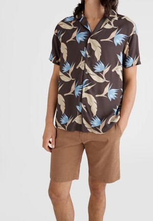 Homme portant un short marron et une chemise à manches courtes à boutons avec un imprimé de feuilles tropicales bleues et beiges, les mains dans les poches.