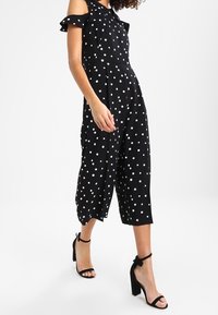 Jumpsuit noir à pois avec des manches dénudées, jambes raccourcies larges et taille ajustée. Porté avec des sandales à talons noires.