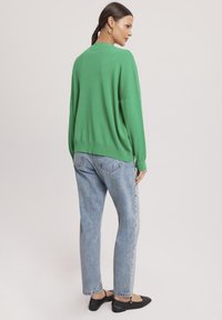 Zielony, oversize sweter z okrągłym dekoltem i opadającymi ramionami, w połączeniu z niebieskimi dżinsami i czarnymi balerinami, o luźnym kroju.