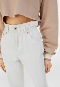 Pantalones de mezclilla blanca de cintura alta con cintura elástica, bolsillos frontales y herrajes en tono cobre, combinados con un sudadera corta beige.