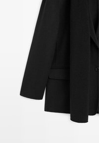 Černý blazer ze směsi vlny se strukturovanými klopami, zapínáním na jeden knoflík a dlouhými rukávy. Tkanina má jemnou strukturu.