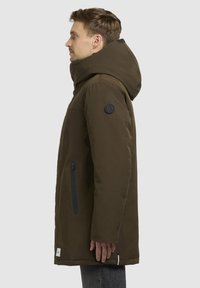 Brun isoleret parka med høj krave, lynlåslomme på siden og et logo-patch på den venstre ærme. Glat tekstur, mellemlang design.