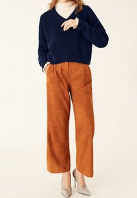 Kobieta ubrana w granatowy sweter dzianinowy, białą bluzkę z falbankami przy mankietach, szerokie brązowe spodnie oraz beżowe czółenka w szpic.