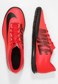 Zapatos de fútbol rojos con parte superior texturizada, swoosh negro de Nike y letras negras audaces. Cuenta con una suela de goma para tracción.