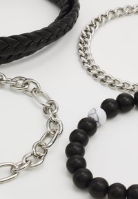 Trois bracelets : une pièce en cuir noir tressé, une chaîne en argent chunky, et un design de perles avec des perles noires et un accent en marbre blanc.