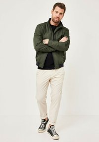 Grüne Bomberjacke mit Reißverschluss, schwarzes Hemd darunter, hellbeige Hosen und schwarze High-Top-Sneaker. Das Model steht mit verschränkten Armen.