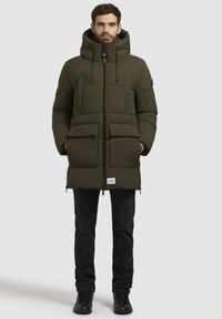 Olivengrøn pufferjakke med høj krave, lynlåslukning, fire forlommer og sorte detaljer. Båret over mørke jeans og sort fodtøj.
