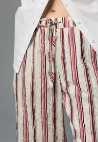 Crème gestreepte broek met rode en zwarte lijnen, gestructureerde stof, losse pasvorm en een gestrikte tailleband, gedeeltelijk zichtbaar onder een wit shirt.