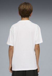 Παιδί με κοντά καστανά μαλλιά που φοράει απλό λευκό μπλουζάκι και μαύρο σορτς, με την πλάτη προς τα έξω, μπροστά σε ένα γκρι διαβαθμισμένο φόντο.