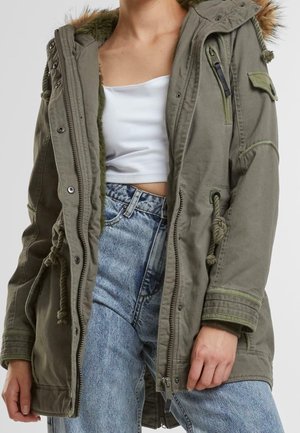 Mujer con parka verde oliva abierta con ribete de piel, top blanco recortado y jeans azul claro de talle alto, con la mano apoyada al costado.