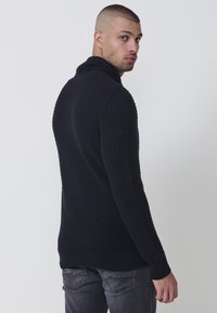 Marineblauer gestrickter Pullover mit einem strukturierten Muster und hohem Kragen. Hat lange Ärmel und einen gerippten Saum. Kombiniert mit dunkelgrauen Jeans.