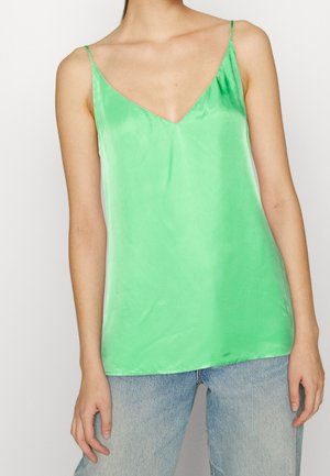 Vrouw die een felgroen satijnen hemdje met spaghettibandjes en een blauwe jeans draagt, staand tegen een effen lichte achtergrond.