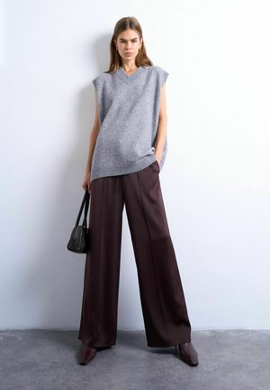 Kvinde iført ærmeløs grå strikket vest, viddebenede brune bukser og brune spidse sko, der holder en sort håndtaske mod en hvid væg.