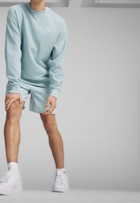 Person klädd i ljusblå Puma-sweatshirt och matchande shorts, vita strumpor och vita sneakers, som håller ett knä och står på en grå golv.