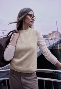Pull en tricot crème avec un col côtelé, superposé sur un haut à manches longues léger, associé à un pantalon marron texturé et un sac marron.