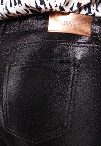 Svarta denimjeans med en glansig yta, med en synlig bakficka och en metallisk kopparfärgad Hugo-etikett ovanför midjan.