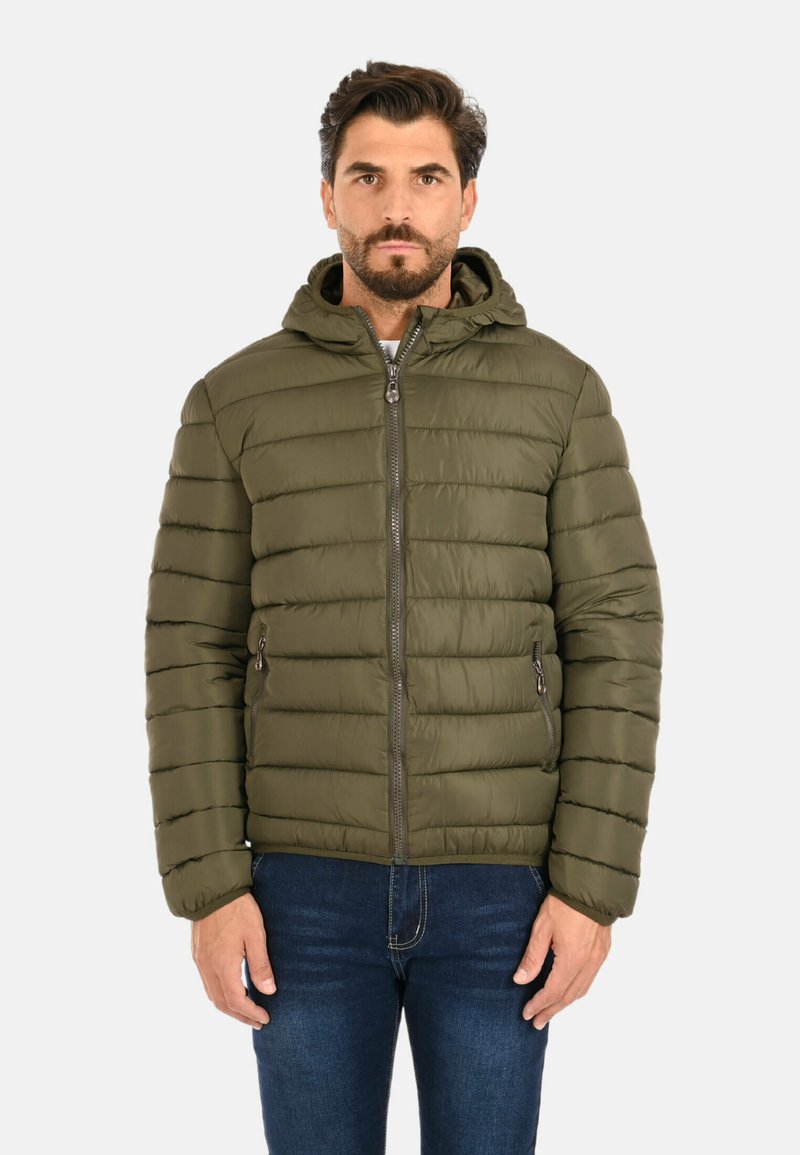 Uomo che indossa una giacca con cappuccio trapuntata verde oliva, chiusa, abbinata a jeans blu scuro, in piedi contro uno sfondo neutro.