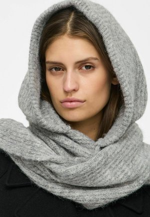 Une femme porte une épaisse écharpe en maille gris clair, enroulée sur sa tête et autour de son cou. Elle porte un manteau noir sur un fond uni.