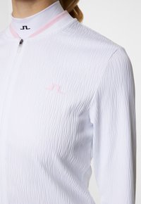 Vit zip-up jacka med texturerad yta, rosa detaljer vid kragen och en subtil logotyp. Materialet verkar lätt och något elastiskt.