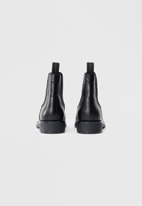 Une paire de bottes Chelsea noires en cuir avec des languettes portant l'inscription "GANT", montrées de l'arrière sur un fond blanc.