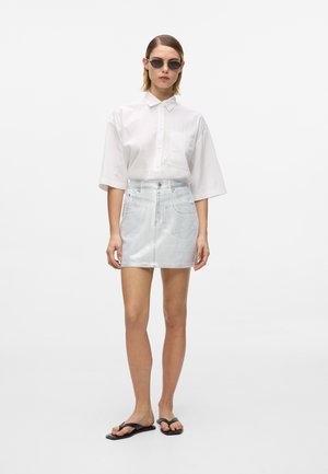 Mujer con gafas de sol negras, camisa blanca de manga corta y oversize, minifalda de mezclilla clara y chanclas negras, de pie contra un fondo blanco.