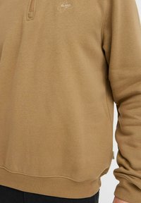 Brown sweatshirt with a half-zip design, featuring a small logo on the chest and ribbed cuffs. Made from a soft, textured fabric.