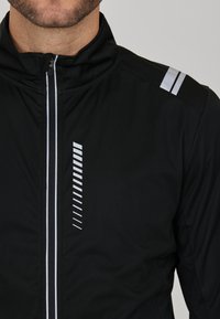 Svart zip-up jacka med reflekterande silvriga ränder på axlarna och vertikala linjer på bröstet. Slät, lätt tygliknande textur.