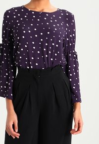 Blusa viola a pois con maniche lunghe e polsini a sbuffo, abbinata a pantaloni neri a vita alta, con pieghe verticali e una cerniera.