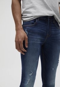 Vaqueros de mezclilla azul desgastados con detalles desgarrados, diseño de cinco bolsillos y costuras en contraste. La tela tiene una textura suave, silueta ajustada.