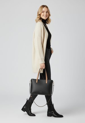 Jeune femme en cardigan beige, haut noir, jean foncé et bottines noires marchant tout en portant un sac à main noir avec des poignées en cuir clair.