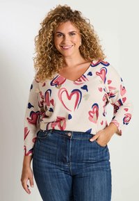 Pull en tricot beige avec un col en V, arborant des motifs de cœurs multicolores avec des accents rose, bleu et or. Associé à un jean bleu foncé.