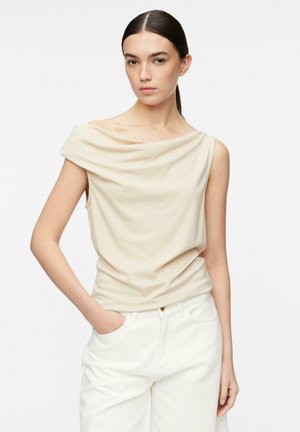 Młoda kobieta o ciemnych włosach, ubrana w beżowy bezrękawnik z drapowaniem i białe spodnie, z jedną ręką w kieszeni, neutralne tło.