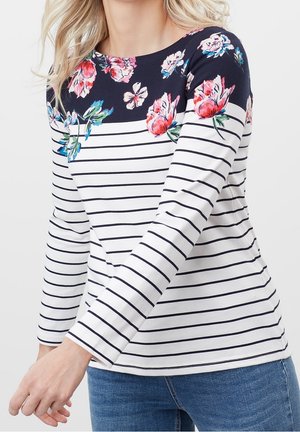 Femme portant une chemise blanche à manches longues avec de fines rayures noires horizontales et une partie supérieure bleu marine décorée de motifs floraux roses et rouges.