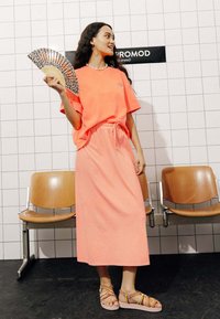 T-shirt surdimensionné en néon orange associé à une jupe maxi assortie, tous deux en tissu doux. Accessoirisé avec un éventail à motifs et des sandales colorées.