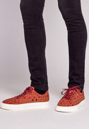 Red patterned sneakers with a floral design, white rubber sole, and red laces, paired with black slim-fit jeans.