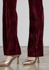Piernas vestidas con pantalones de terciopelo rojo oscuro texturizados, combinados con brillantes zapatos de tacón alto dorados, de pie sobre un suelo pulido.
