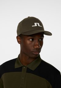 Olivgrön keps med platt skärm, med en framträdande vit logga. Tyget ser slätt ut med strukturerade paneler. Svart polo-tröja under.