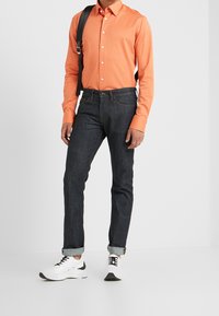 Camisa de manga larga naranja con botones, jeans de mezclilla oscura y zapatillas blancas con acentos negros. La camisa tiene un diseño metido por dentro y mangas enrolladas.