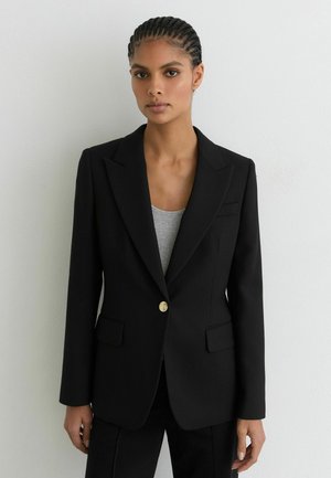 Vrouw met gevlochten haar draagt een zwarte gestructureerde blazer met gouden knopen over een grijze top, staand tegen een effen witte muur.