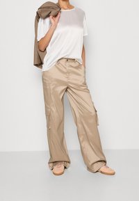 Beżowe, lśniące, szerokie spodnie cargo z dużymi kieszeniami, połączone z gładką białą koszulką. Brązowa kurtka jest zawieszona na ramieniu.