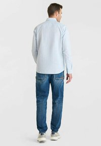 Man die met zijn rug naar de kijker staat, gekleed in een lichtblauw gestreept shirt, blauwe spijkerbroek met achterzakken en witte sneakers, tegen een effen achtergrond.