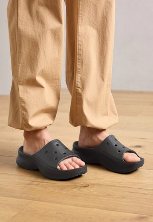 Personne portant un pantalon beige et des sabots noirs à enfiler avec des trous de ventilation, debout sur un sol en bois.