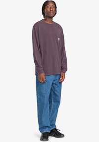 Long-sleeve, dark purple t-shirt with a chest pocket, paired with light blue, loose-fit jeans. Model wearing black sneakers.