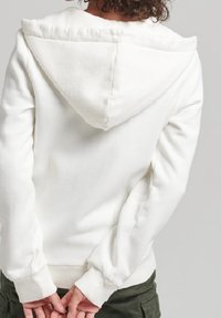 Sudadera blanca con capucha amplia, puños y dobladillo acanalados. Tejido suave y texturizado; la vista trasera muestra líneas limpias y un diseño minimalista.