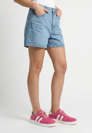 Person iført lyseblå denimshorts og klare pink sneakers med hvide striber, stående mod en ensfarvet hvid baggrund.