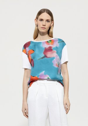 Satin-Mix-Oberteil mit türkisfarbener Basis und mehrfarbigen Blumenmustern; kurze weiße Ärmel und lockere Passform. Kombiniert mit weißen Hosen.