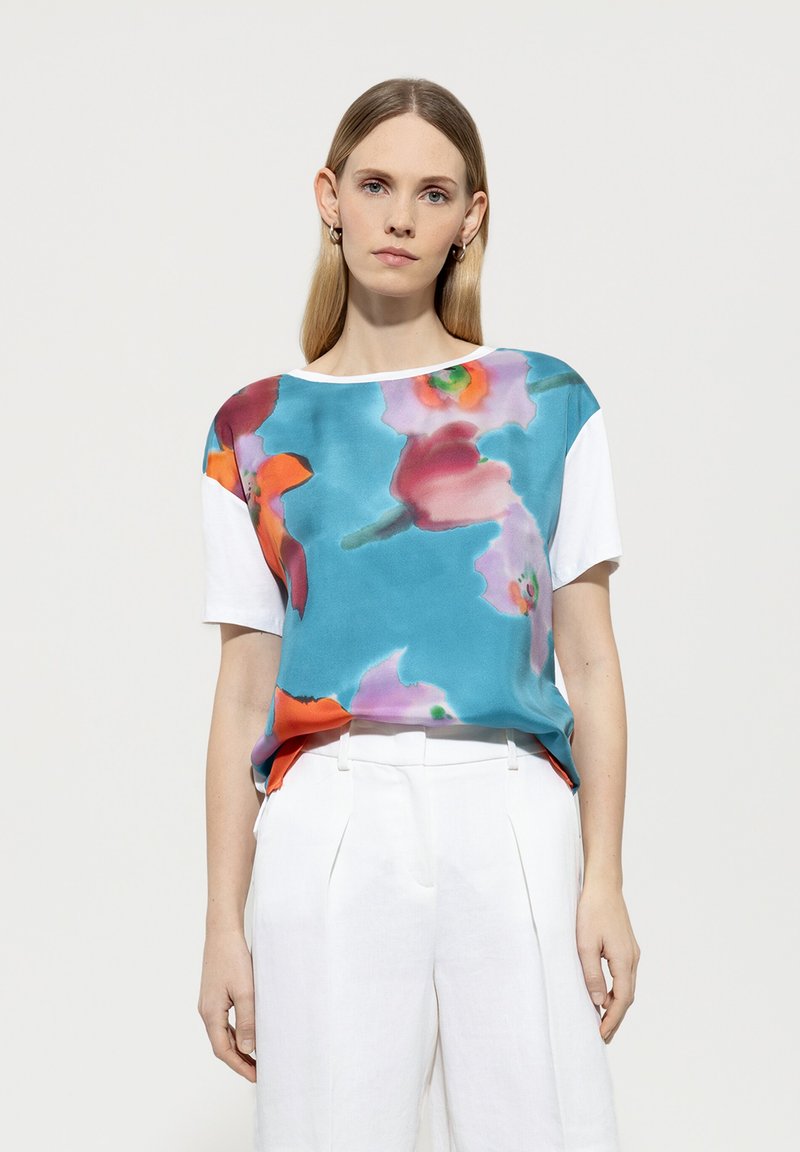 Satin-Mix-Oberteil mit türkisfarbener Basis und mehrfarbigen Blumenmustern; kurze weiße Ärmel und lockere Passform. Kombiniert mit weißen Hosen.